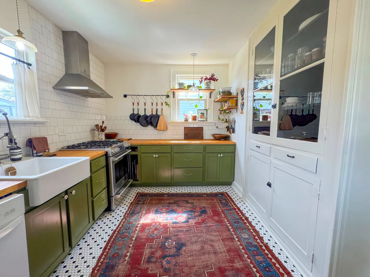 Kitchen with green cabinets, a stainless range and hood, a farmhouse sink, a glass-front cabinet, and a red Persian-style rug.