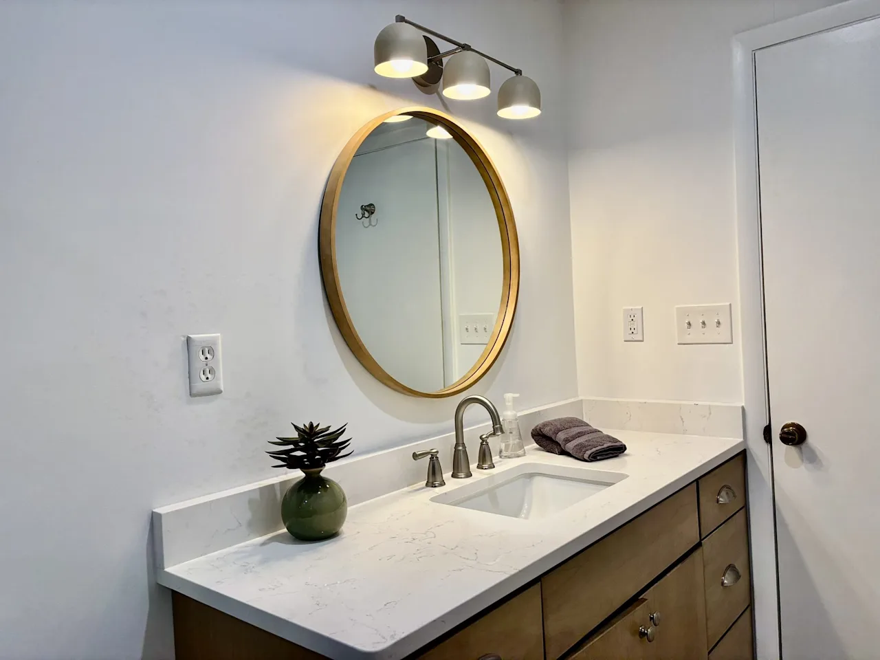 Bathroom vanity with a round gold-framed mirror, quartz counter, and a modern wall sconce on a white wall.