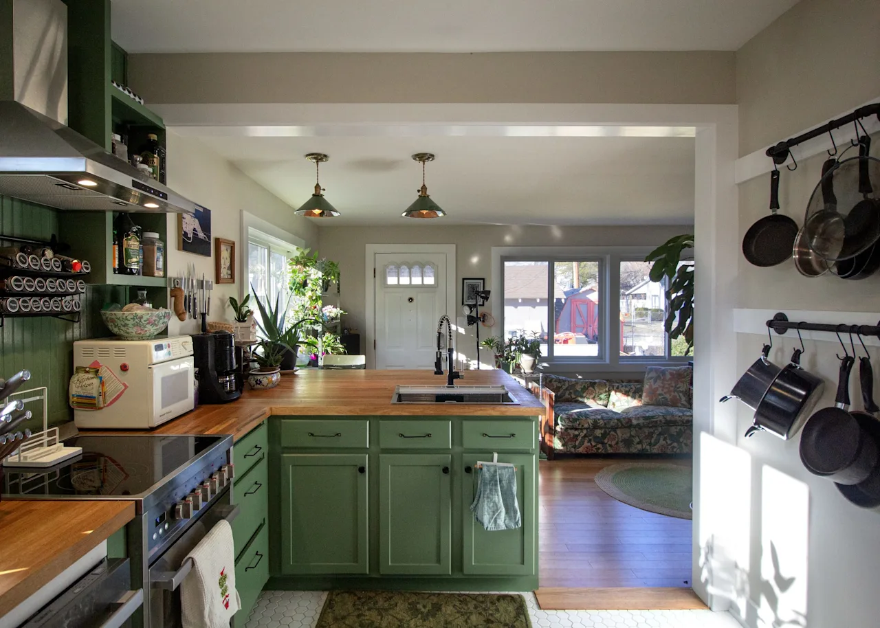 Open-concept kitchen with green lower cabinets, a hanging pot rack, open shelving, and a view through to the living area.