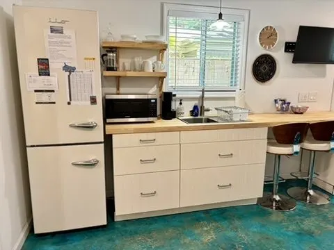 Kitchenette with a cream retro refrigerator, open shelving, a butcher-block counter, and bar stools on the blue concrete floor.