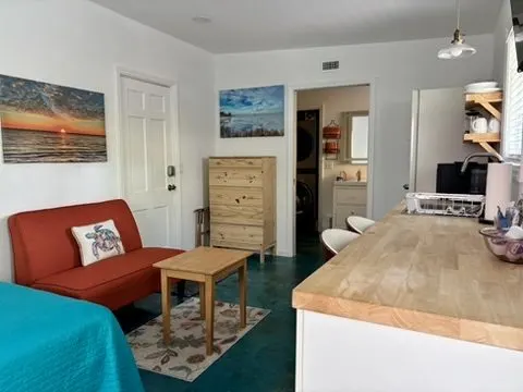 Sitting area with an orange loveseat, a wood coffee table, coastal artwork, and a butcher-block bar on the blue concrete floor.