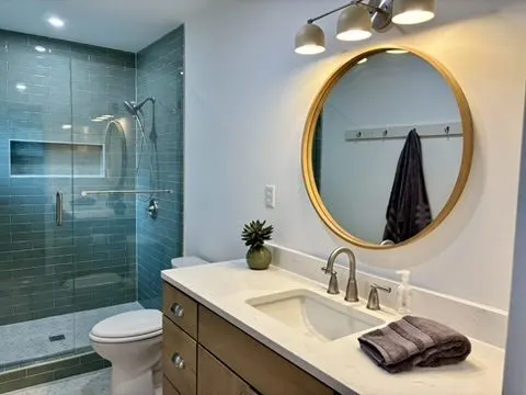 Renovated bathroom with a glass walk-in shower in green tile, a round gold-framed mirror, and a wood vanity with quartz counter.