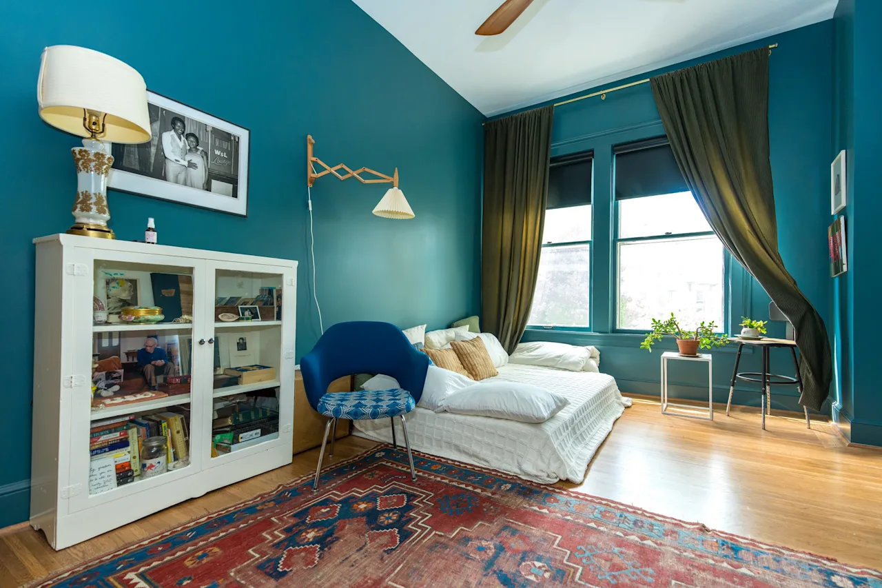 Teal-walled bedroom with a white glass-front cabinet, an upholstered chair, a patterned rug, and a dormer window.