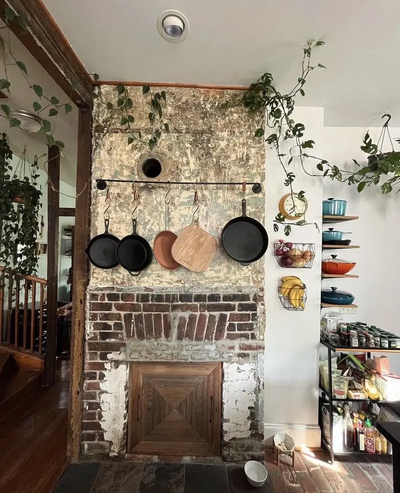 Living space with an exposed brick chimney, cast-iron pans hanging on a rail, trailing plants, and open shelving.