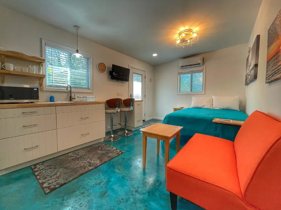 Studio apartment interior with a blue-stained concrete floor, white kitchenette cabinetry, a bright orange sofa, and a bed.