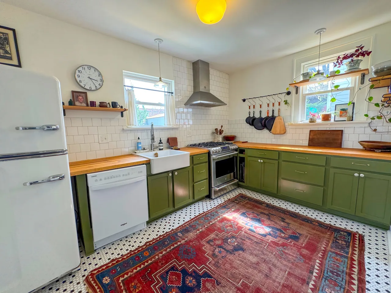 Renovated kitchen with olive-green cabinets, butcher-block counters, white subway tile, a retro fridge, and a red patterned rug on penny-tile floor.