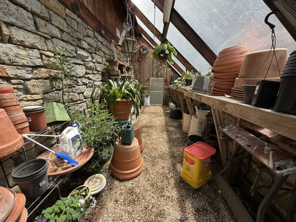Greenhouse potting area along a stone wall, lined with terracotta pots, potted plants, and gardening supplies on a wooden bench.