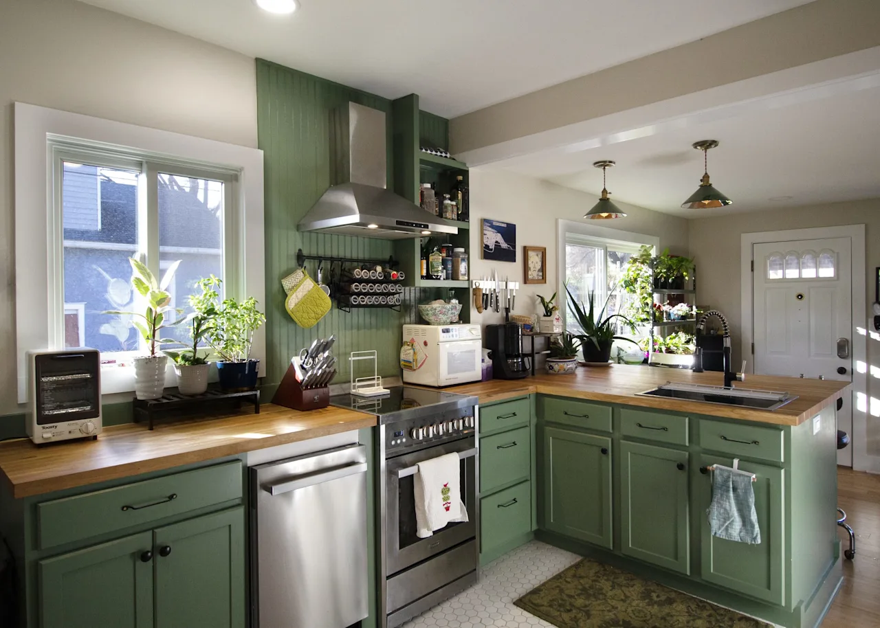 Green-cabinet kitchen with butcher-block countertops, stainless range and dishwasher, and a front door with plants on the sill.