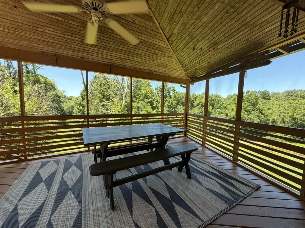 Covered wraparound deck with a timber ceiling and ceiling fan, a black picnic table on a geometric painted rug, overlooking green treetops.