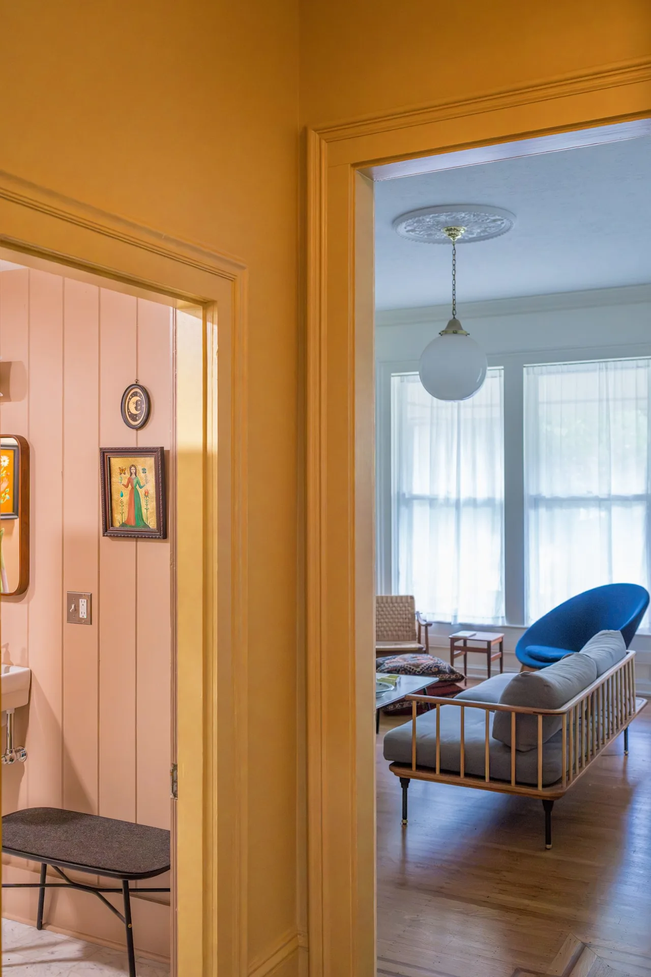 View from a peach-painted room through a doorway into a sitting area with mid-century chairs and a hanging paper lantern.