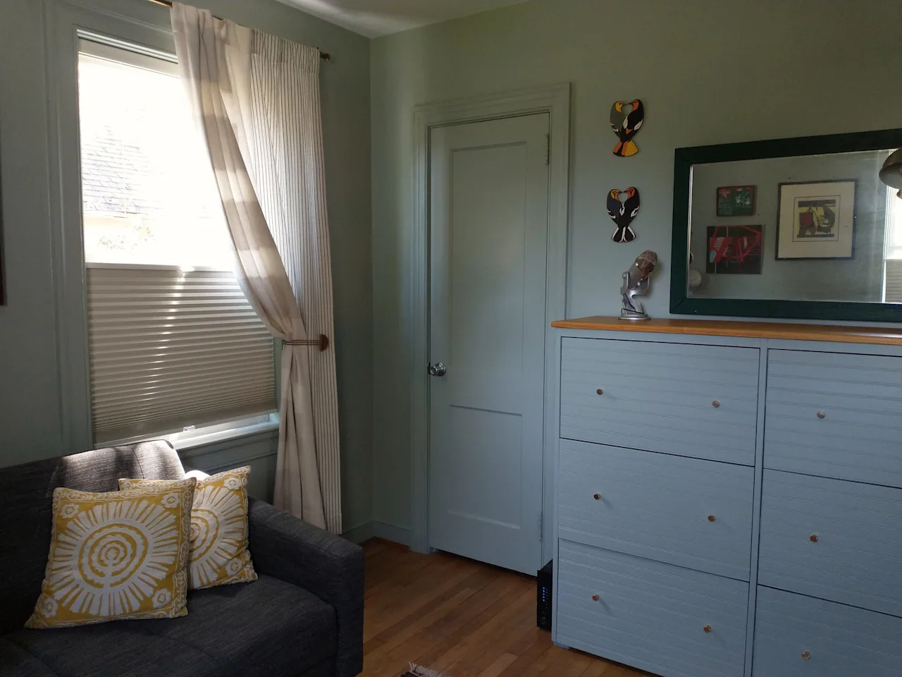 Bedroom corner with a pale-blue dresser, framed art, and a curtained window beside a sofa.