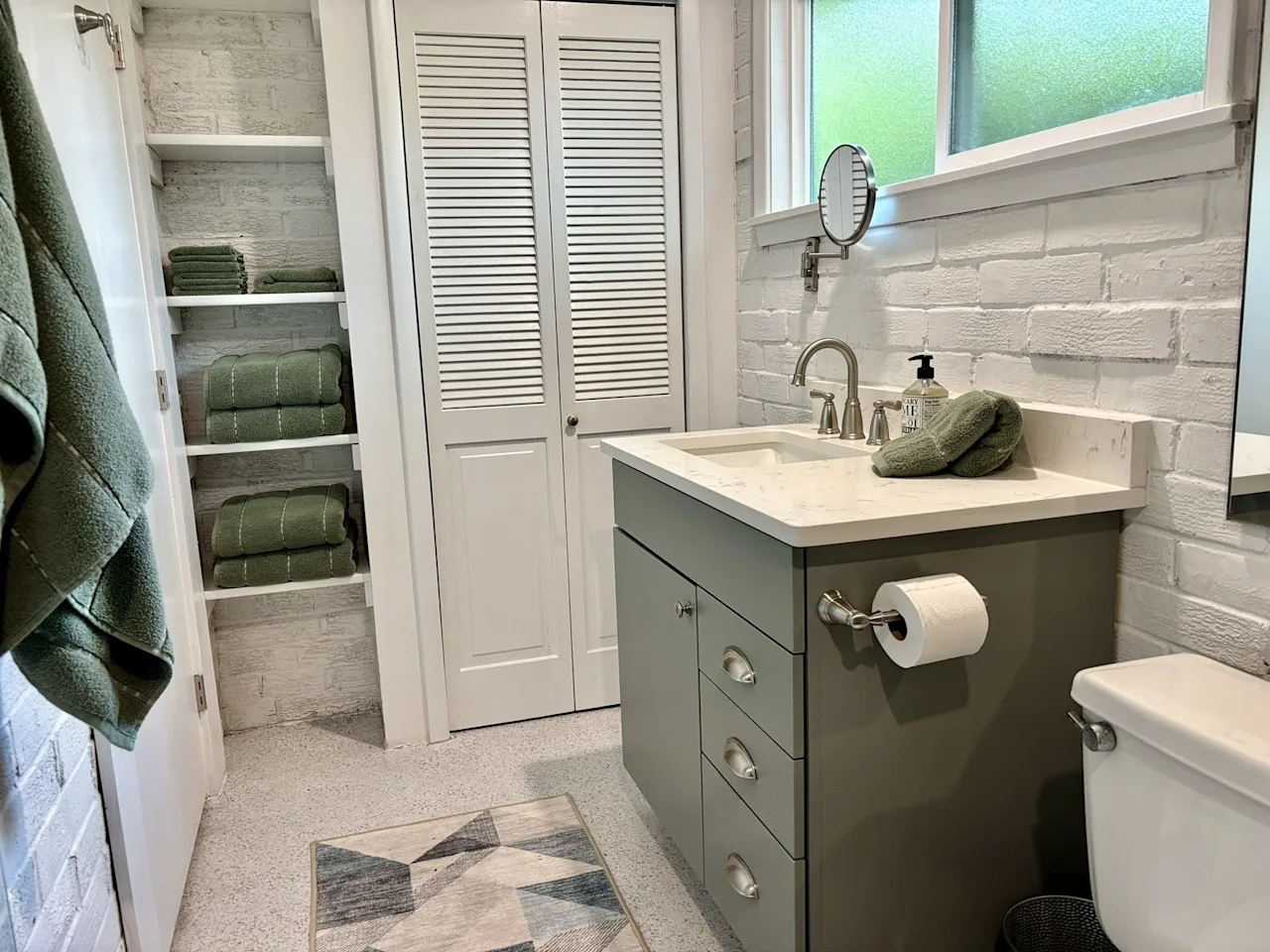 Bathroom with a louvered linen closet, folded green towels, white brick walls, and a wood vanity.