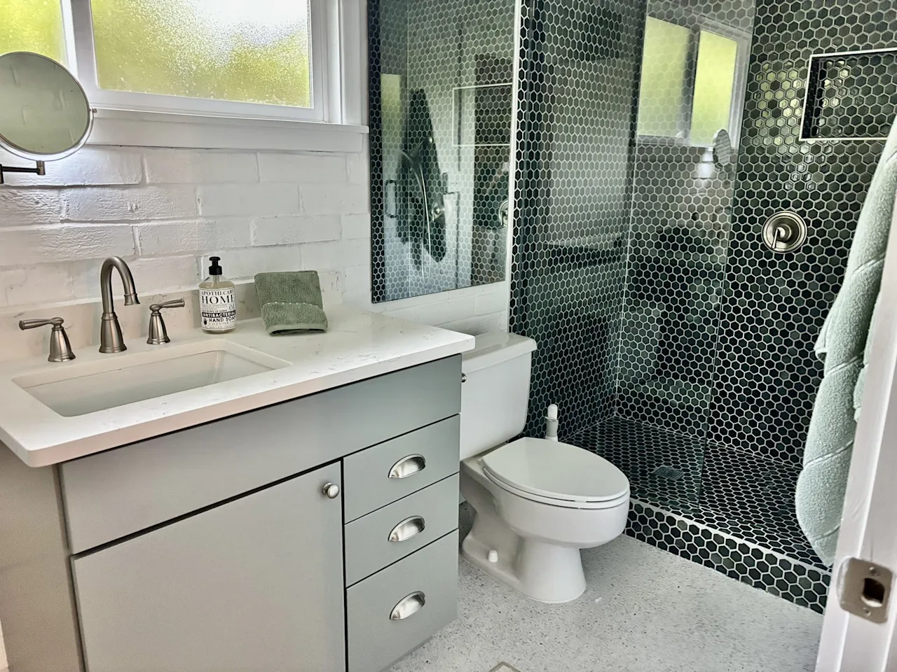 Bathroom with a dark-green penny-tile shower, white vanity, and a frosted window casting green light.