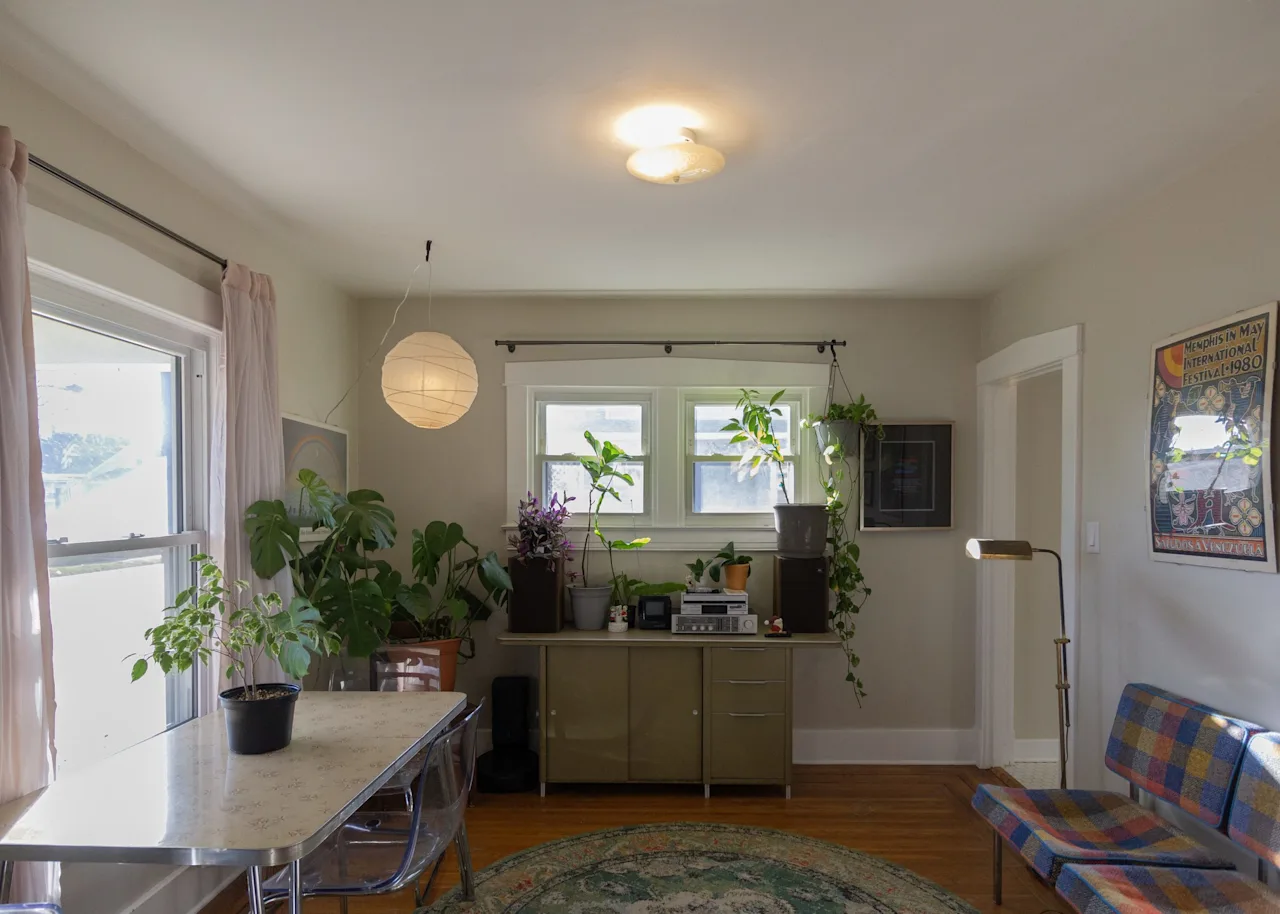 Bright dining and sitting area with a vintage metal table, abundant potted plants, and a plaid loveseat by the windows.