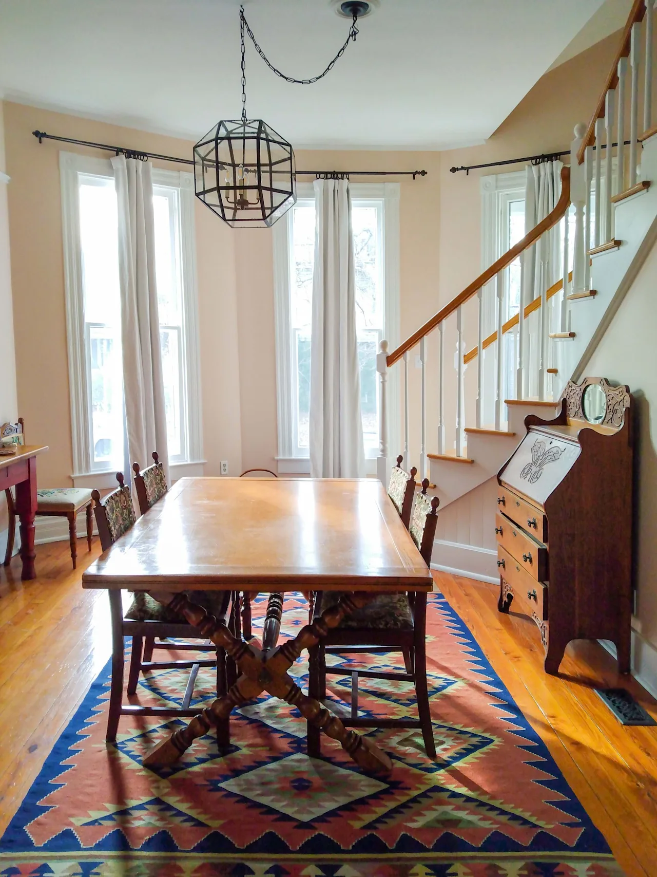 Dining room with a wood table and chairs on a colorful geometric rug, a caged pendant light, tall windows, and a staircase.