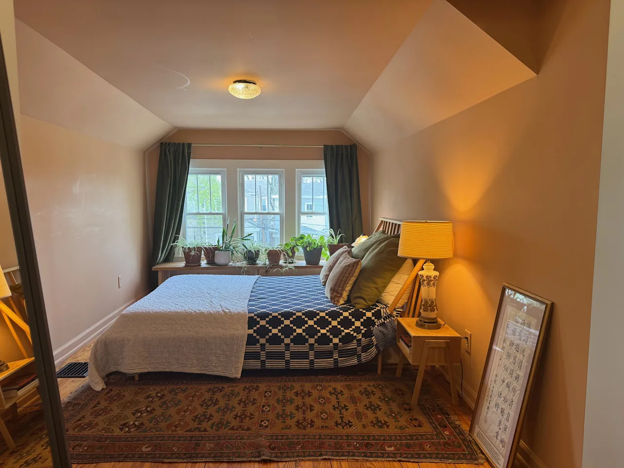 Bedroom under a sloped ceiling with a patterned bedspread, potted plants on the windowsill, and a bedside lamp.