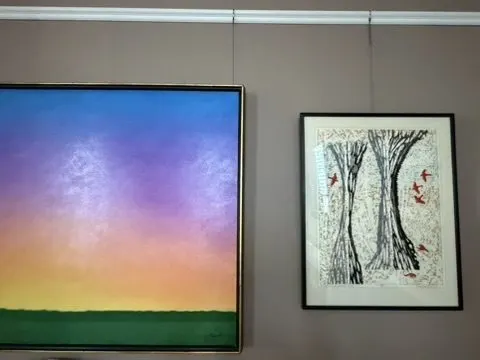 Gallery wall with a colorful sunset landscape painting beside a framed tree artwork on a grey wall.