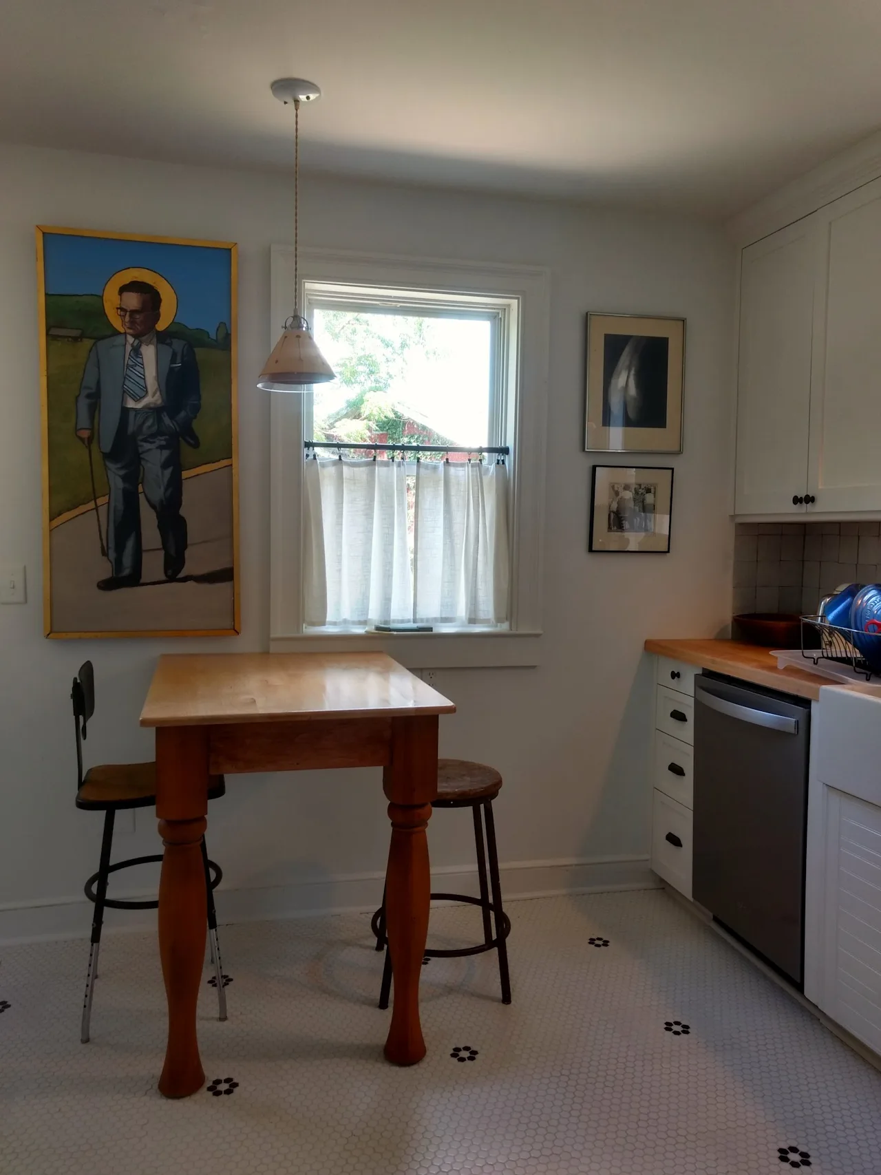 Breakfast nook with a wood table and stools, a framed portrait, a cafe-curtained window, and a penny-tile floor.