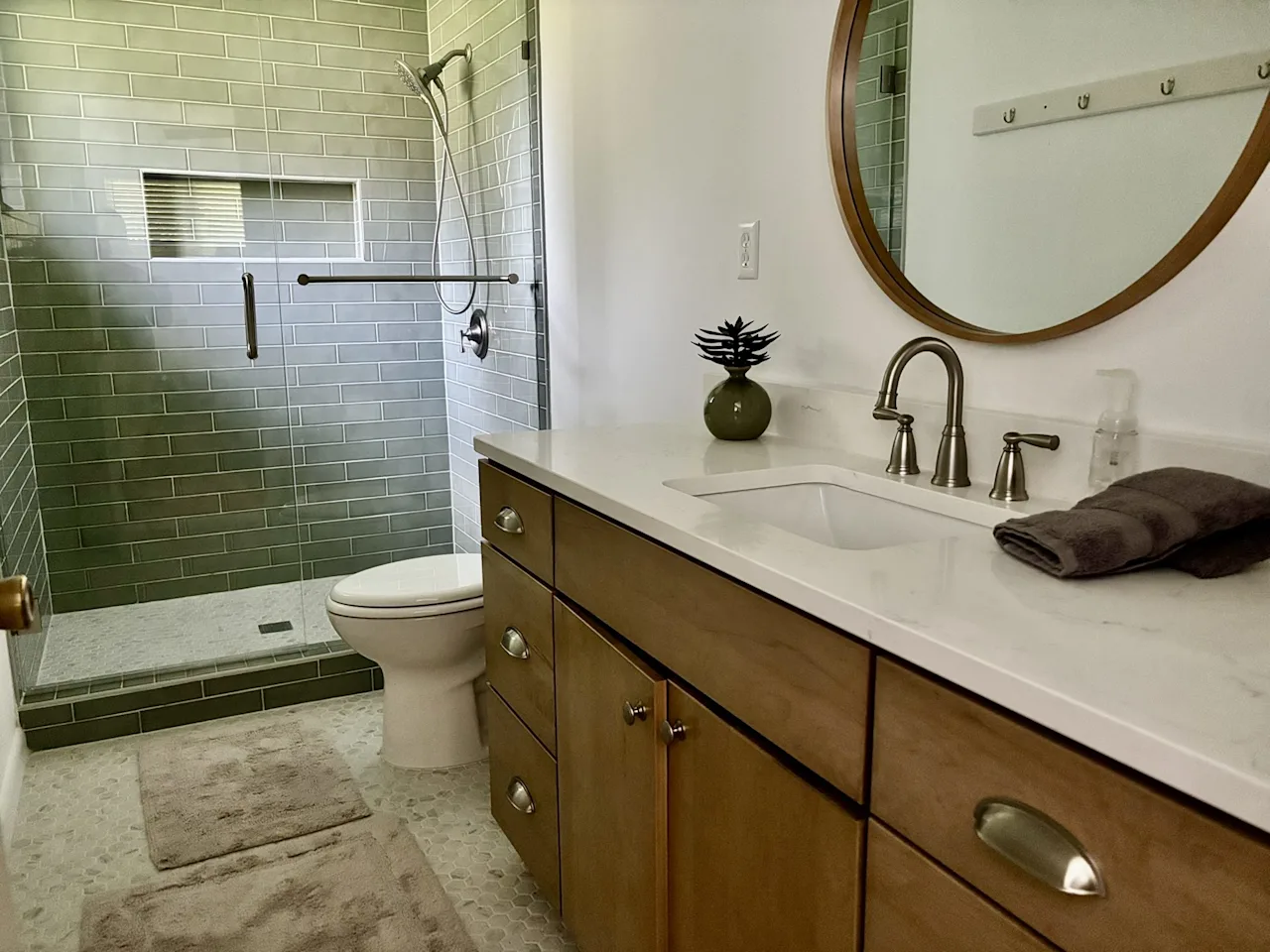 Bathroom with a green subway-tile shower, wood vanity, round mirror, and travertine flooring.