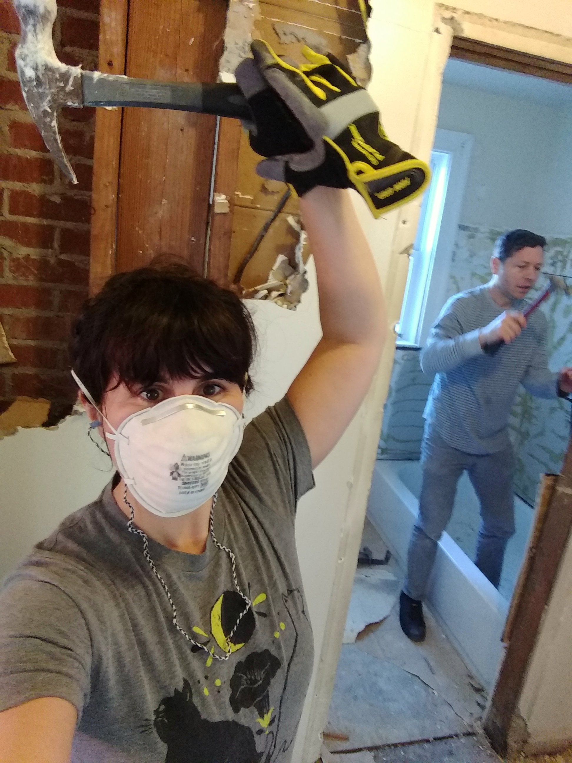 Alea stands with hammer and helper Burton stands in the bathroom, both knocking down walls with hammers.