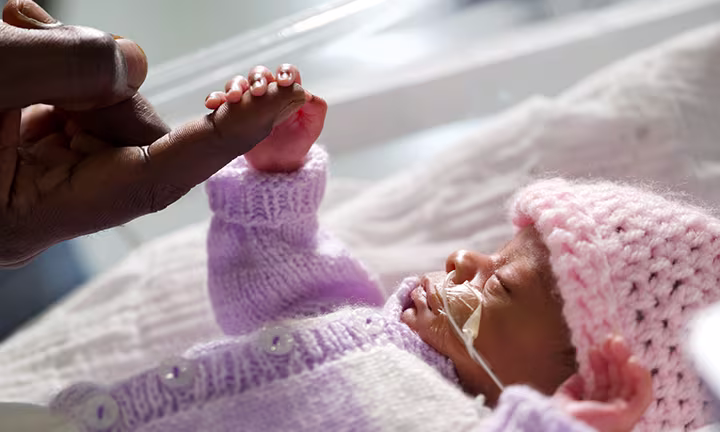 Bébé prématuré en couveuse tenant la main de son parent, portant un bonnet rose tricoté