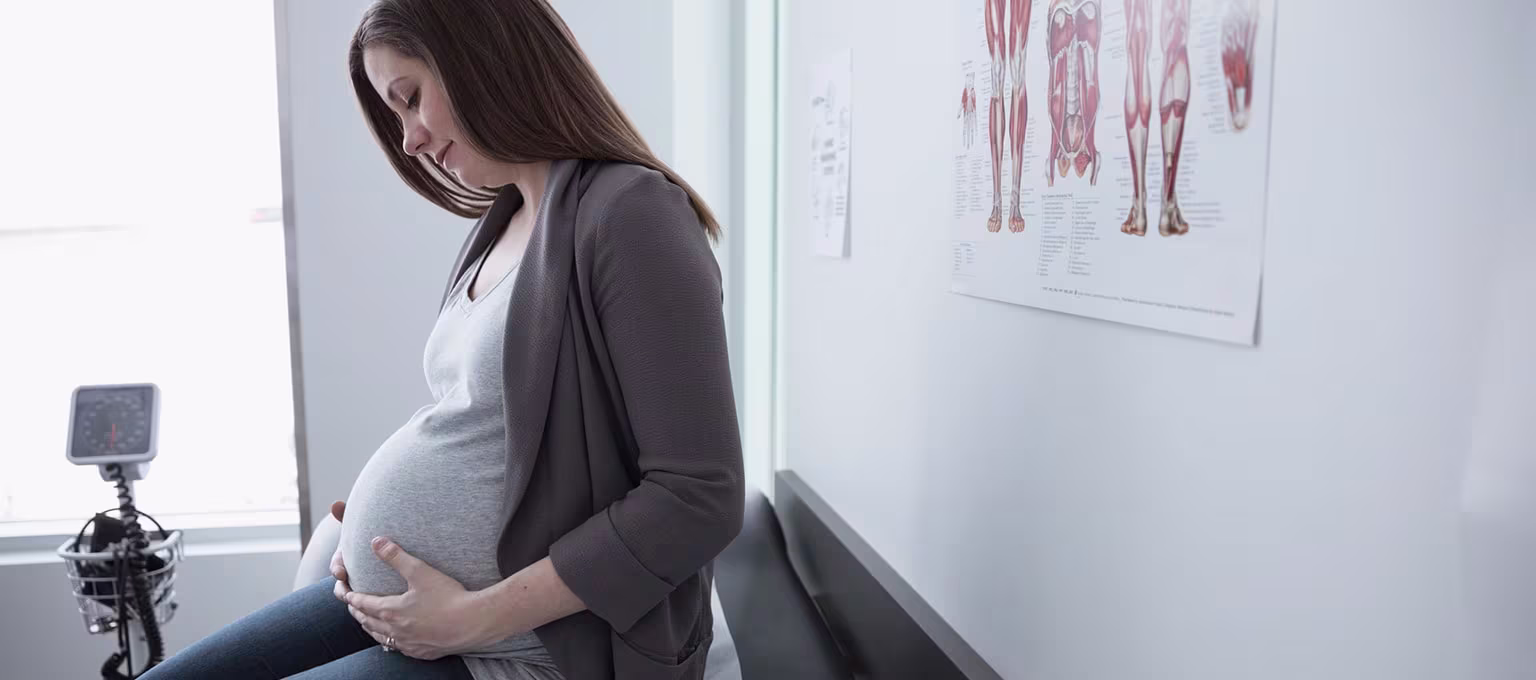 Pregnant woman sitting on a doctor's exam table, holding her belly, possibly preparing for an epidural during labour.