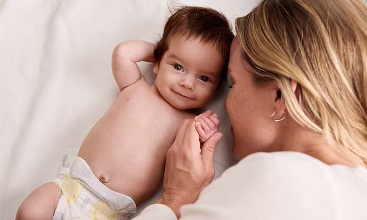 Mama i uśmiechnięte niemowlę leżące twarzą w twarz na miękkim kocyku, dziecko w pieluszce trzyma ją za palec - Obietnica Bezpieczeństwa Pampers