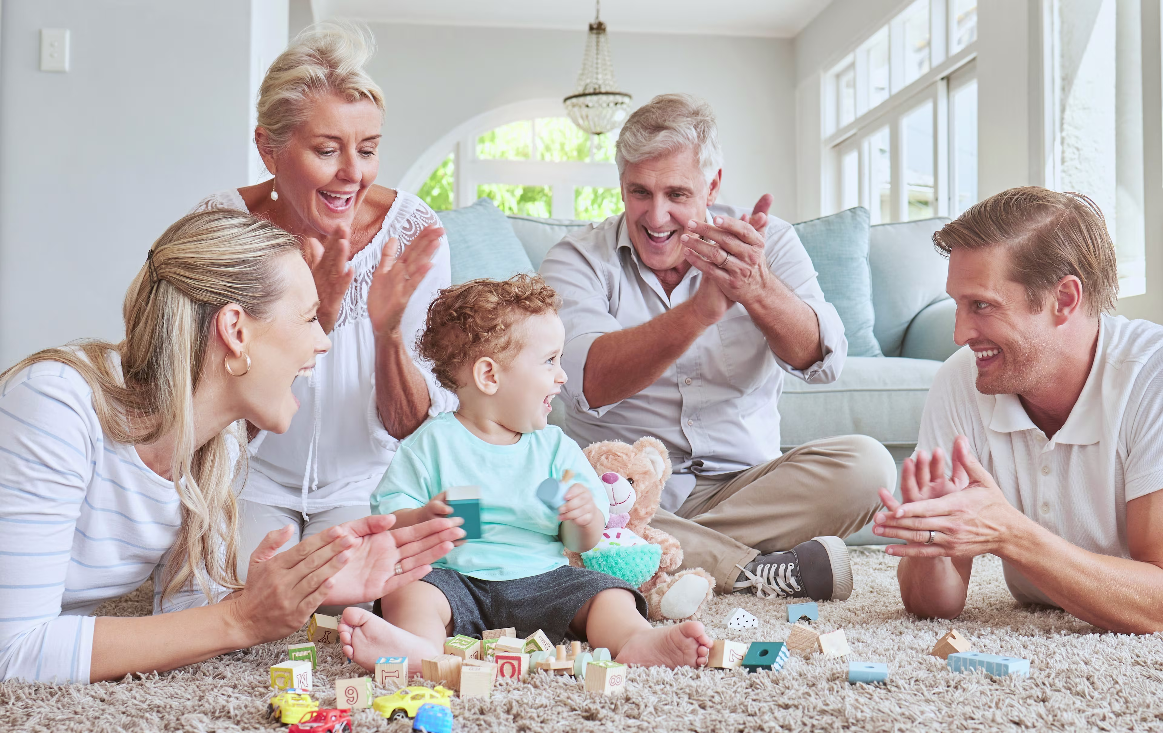 A smiling toddler sits on a living room carpet playing with wooden building blocks as his parents and grandparents gather around clapping and cheering with joy.