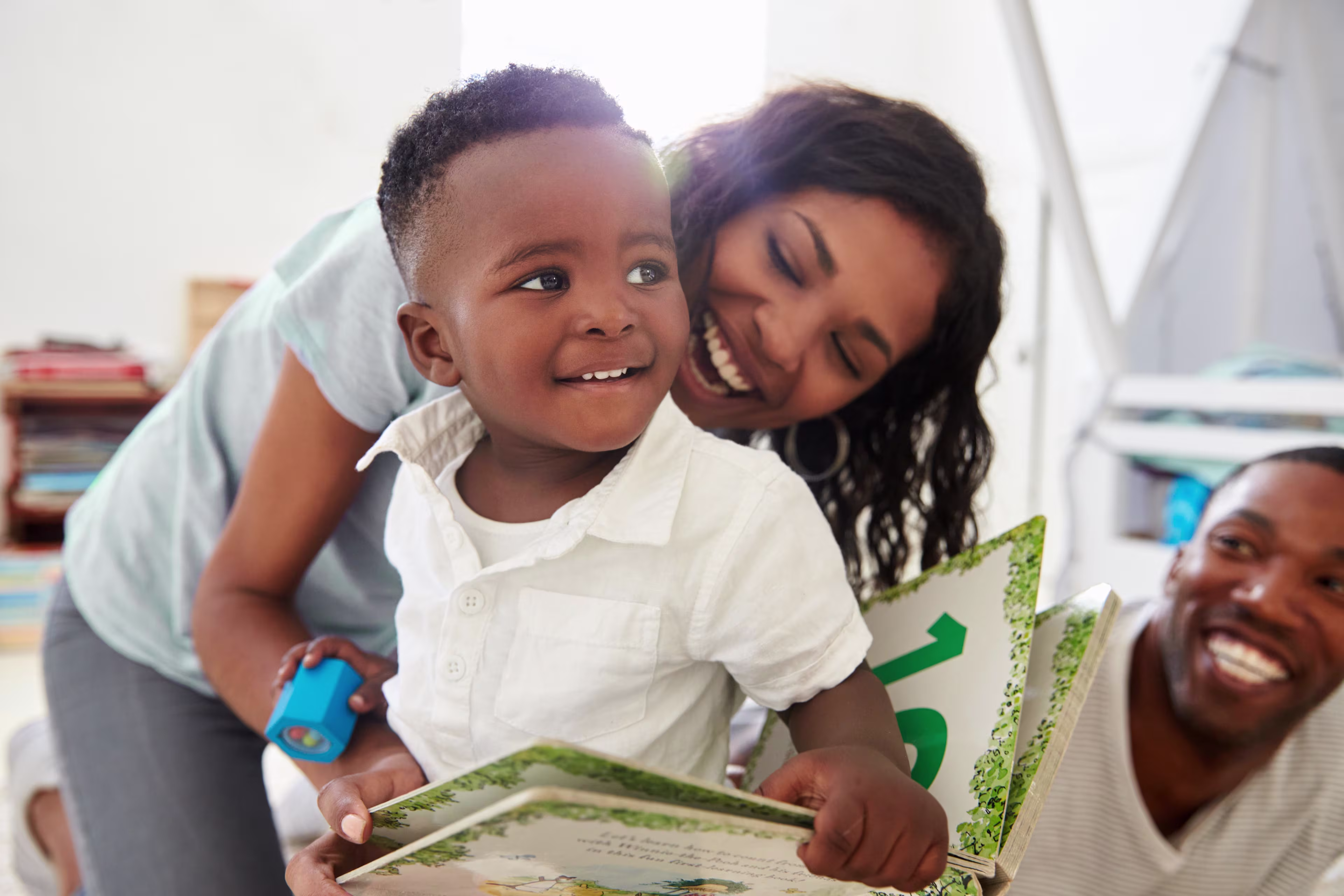 A smiling young Black boy holds an open picture book while his happy parents laugh and look on in a bright room.