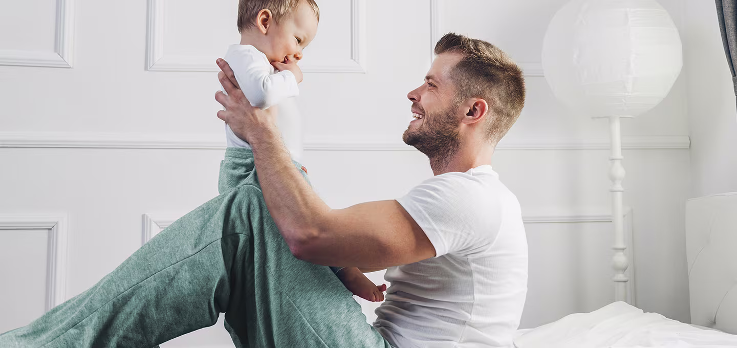 Smiling adult sits on the floor lifting a happy baby who is facing them, showing playful interaction and bonding during simple baby games.
