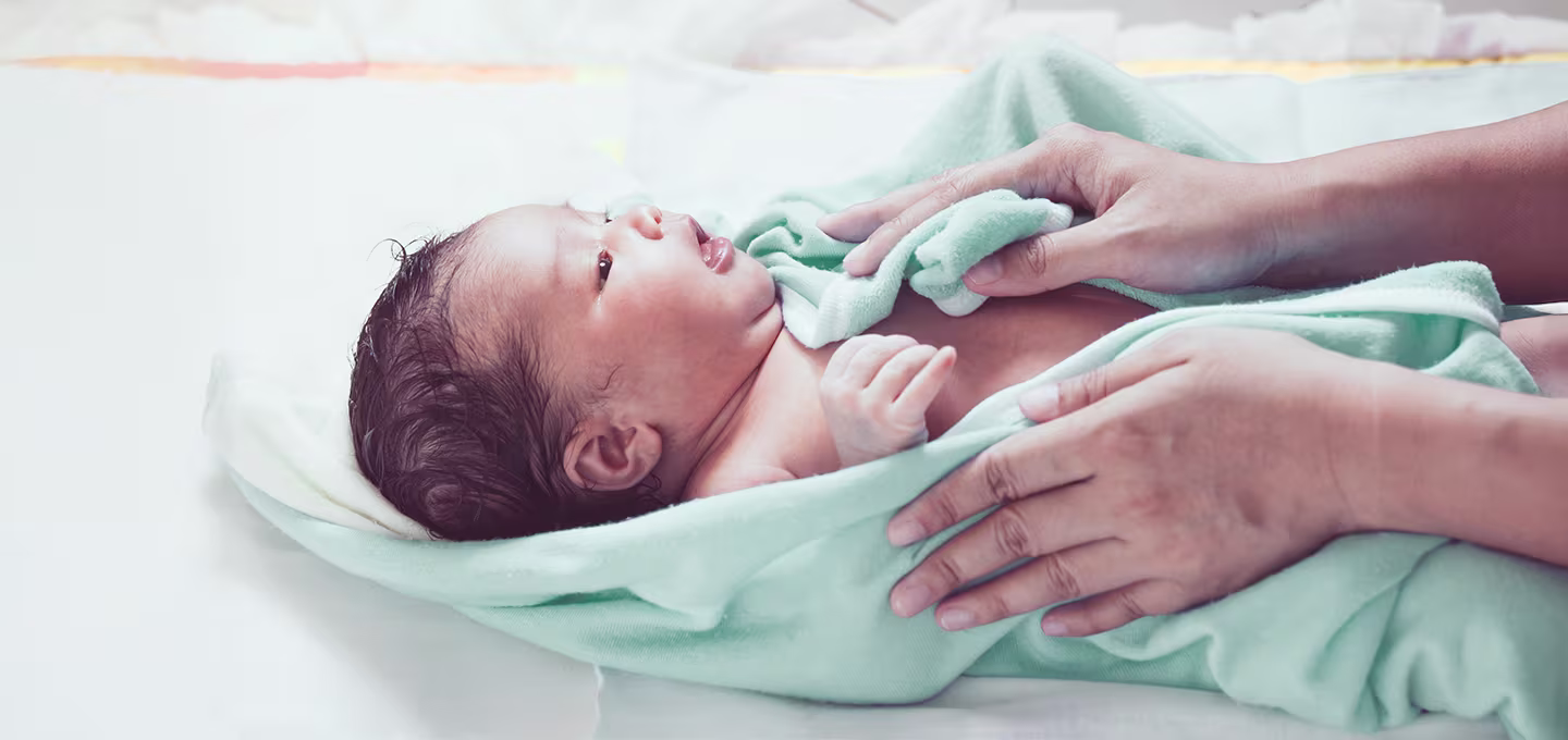 Photo d’un nouveau-né enveloppé dans une serviette bleue après le bain, allongé sur un plan à langer pendant qu’un adulte le sèche et le soutient délicatement. 