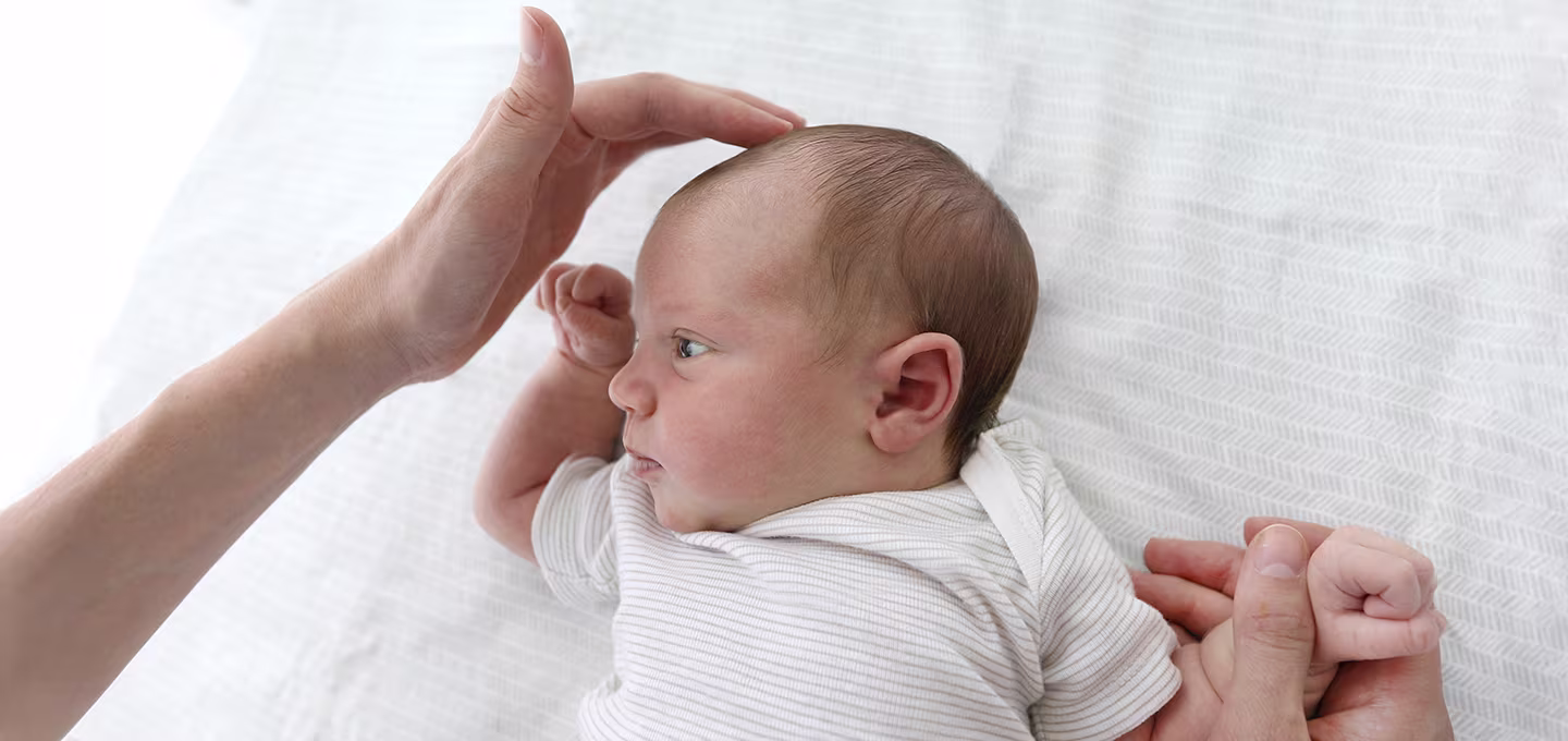 Adult hands gently touching a newborn’s head while the baby lies on a bed, highlighting the soft spot on the top of the baby’s head.