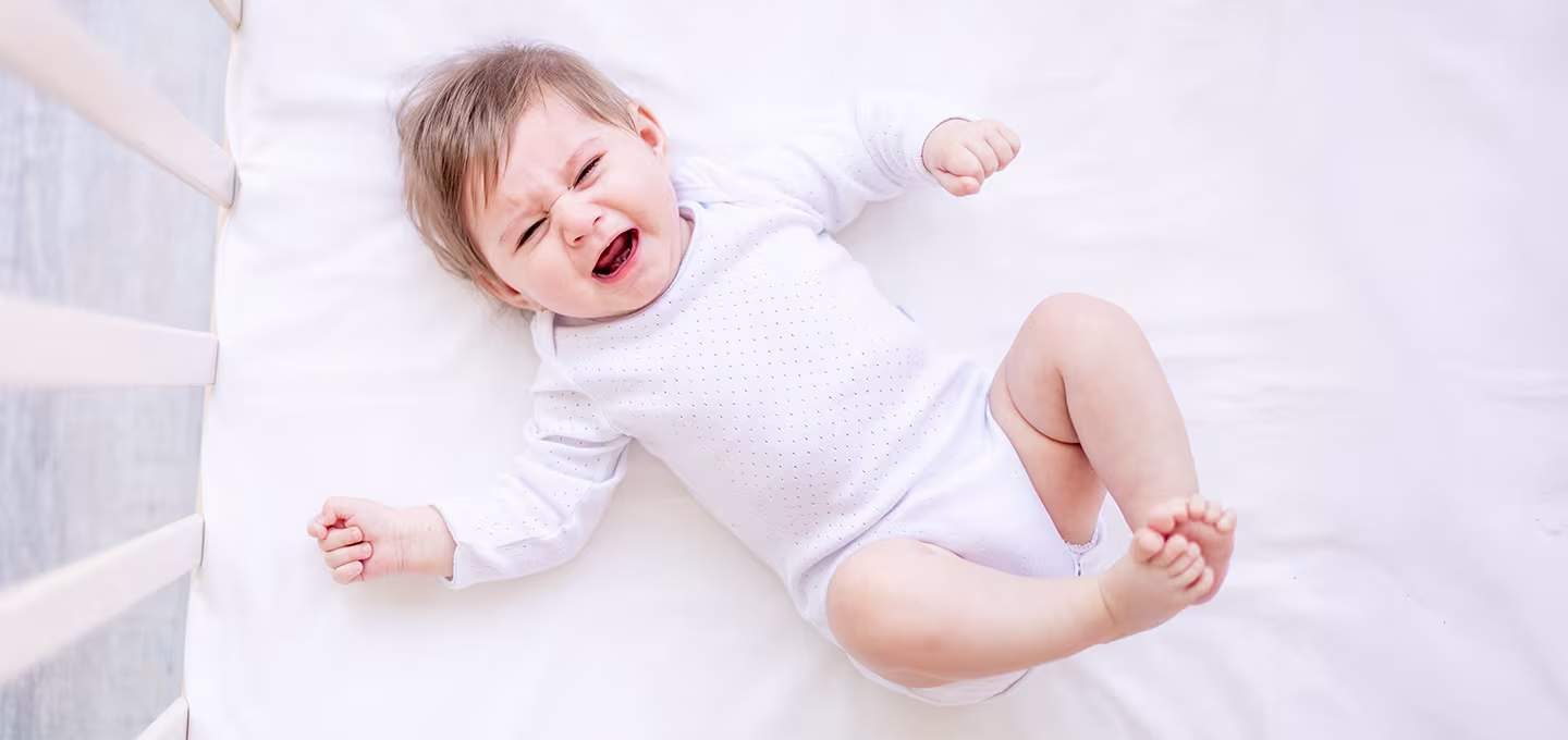 Bebé acostado en cuna llorando con expresión de malestar, piernas y brazos tensos, ilustrando síntomas típicos de cólico infantil.