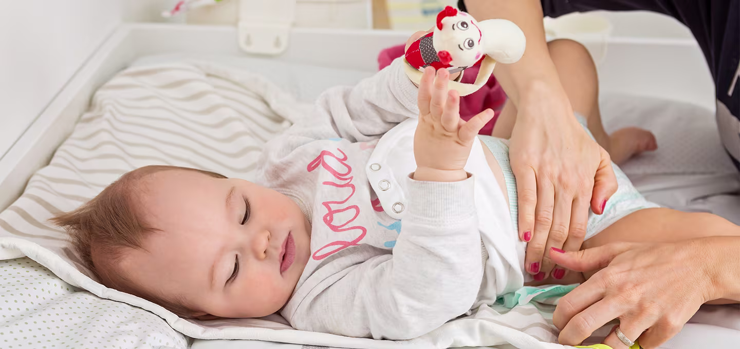 Ein Baby liegt auf einer Wickelunterlage, hält ein Spielzeug, während die Bezugsperson die Windel wechselt