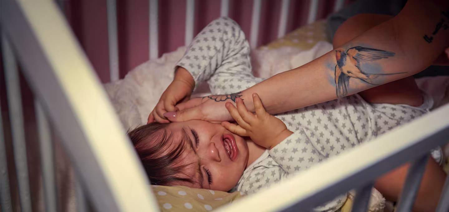Bébé allongé dans un lit pleure pendant qu’un adulte tente de le calmer en le touchant doucement.
