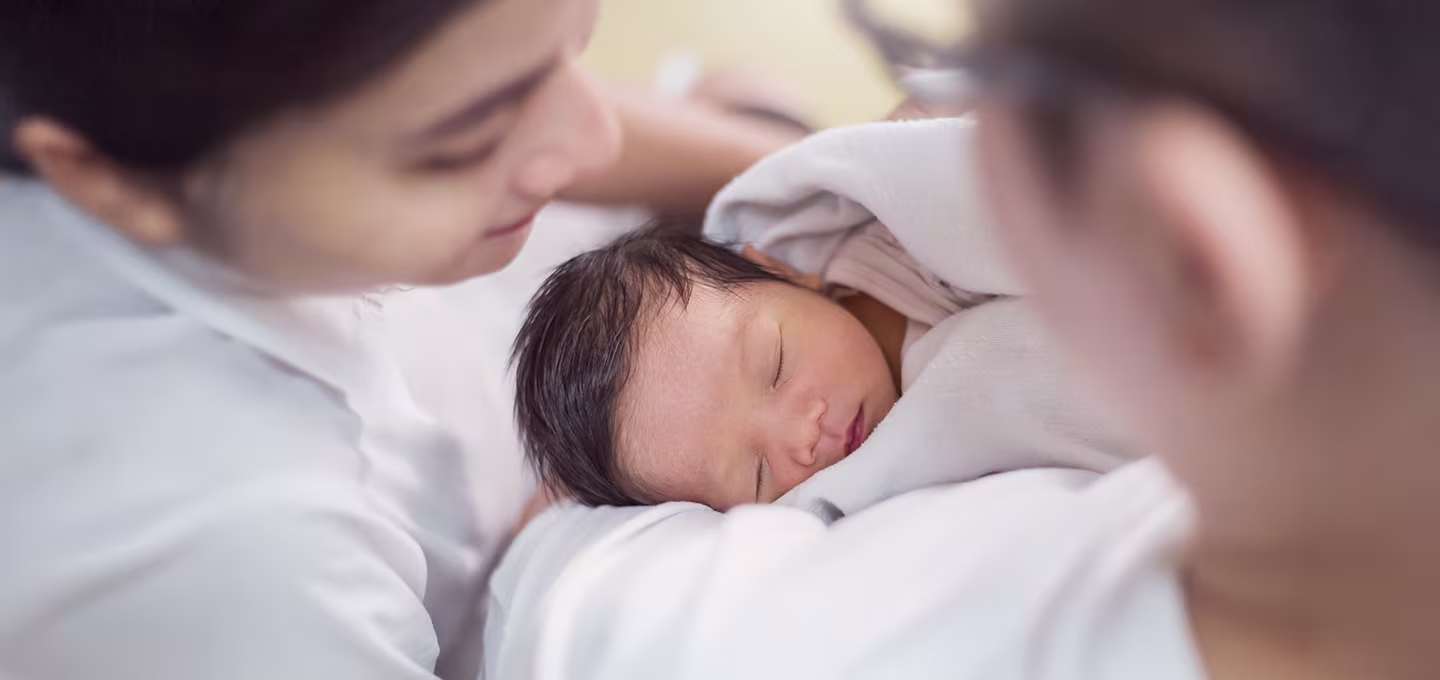 A newborn wrapped in a blanket rests calmly in a caregiver’s arms, showing healthy skin tone as adults gently observe for signs of jaundice.