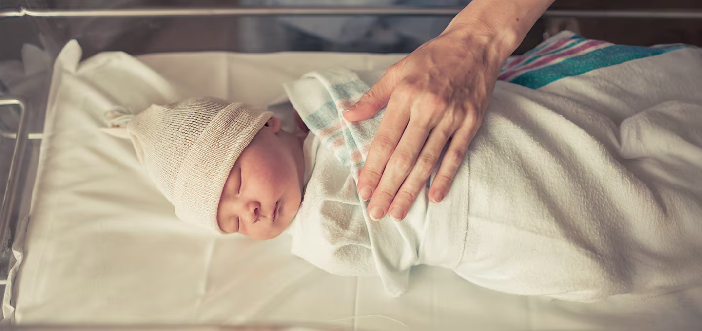 Foto eines neugeborenen Babys, das im Krankenhausbett in eine Decke gewickelt ist, während eine Hand schützend auf seiner Brust liegt.