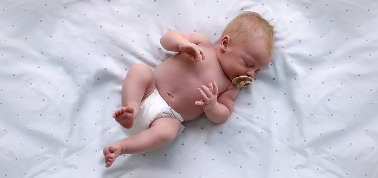 Baby wearing a diaper sleeps on a light polka dot sheet with a dummy in their mouth.