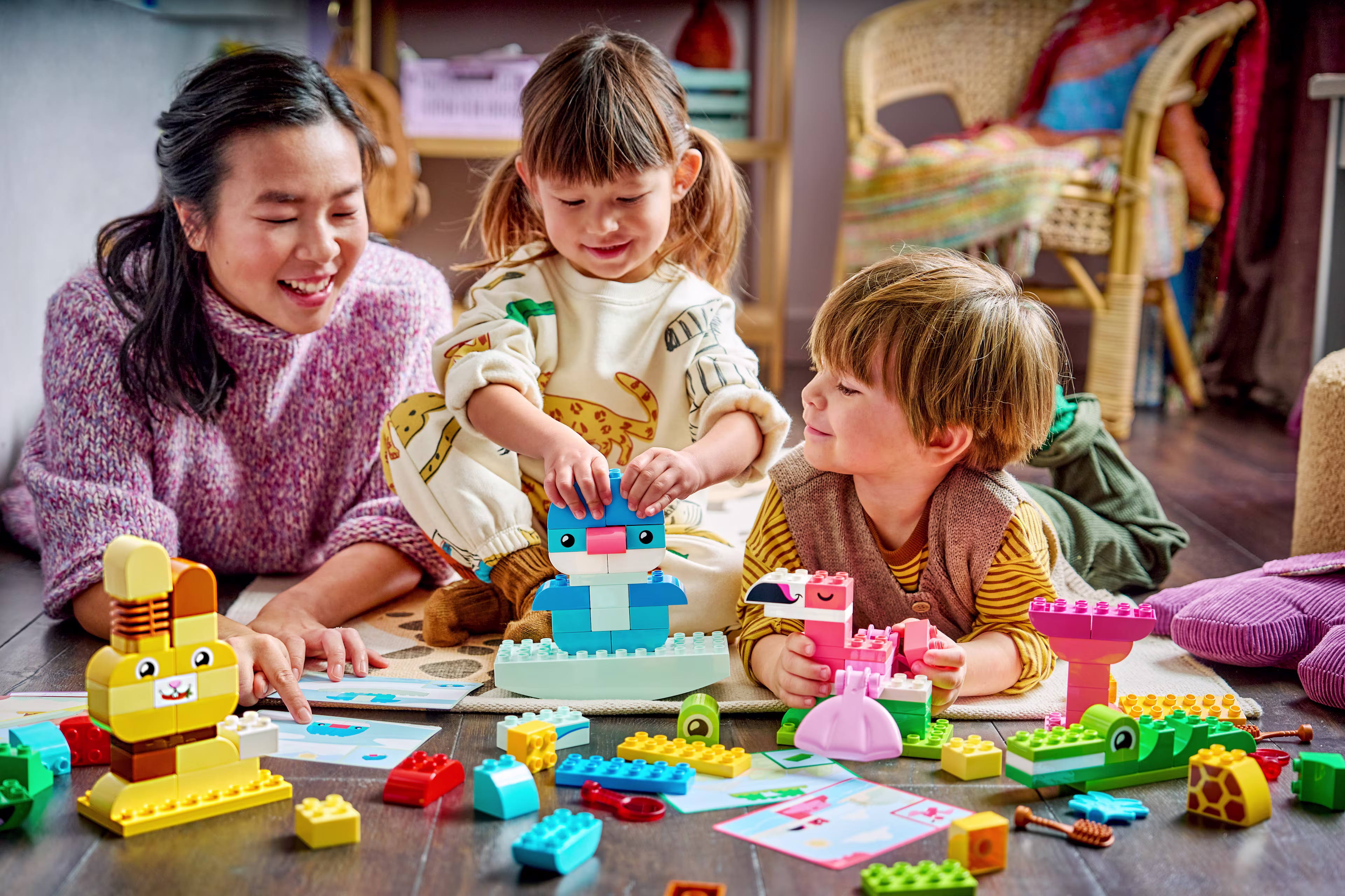 A smiling mother and two young children sit together on a rug, building colorful LEGO DUPLO toy animals from large building blocks.