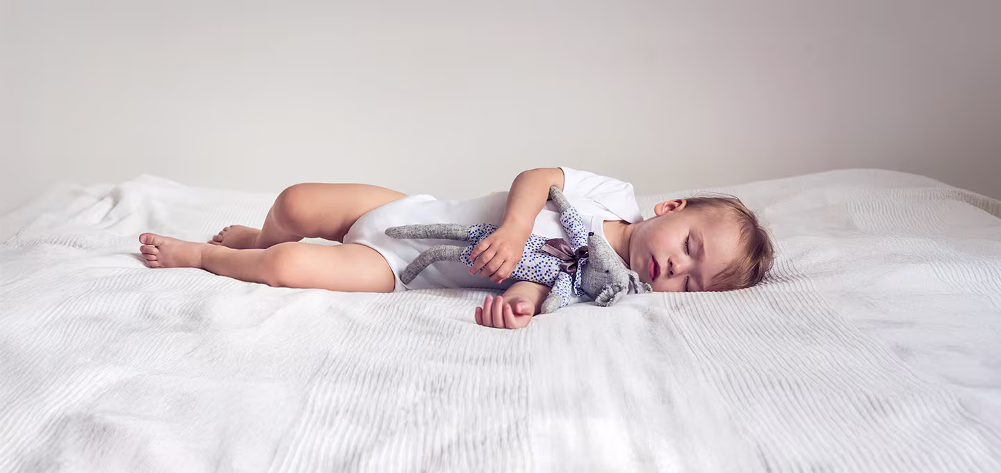 Bébé endormi allongé sur un lit tenant un doudou, illustrant une sieste calme et des horaires de sieste sains pour nourrissons.