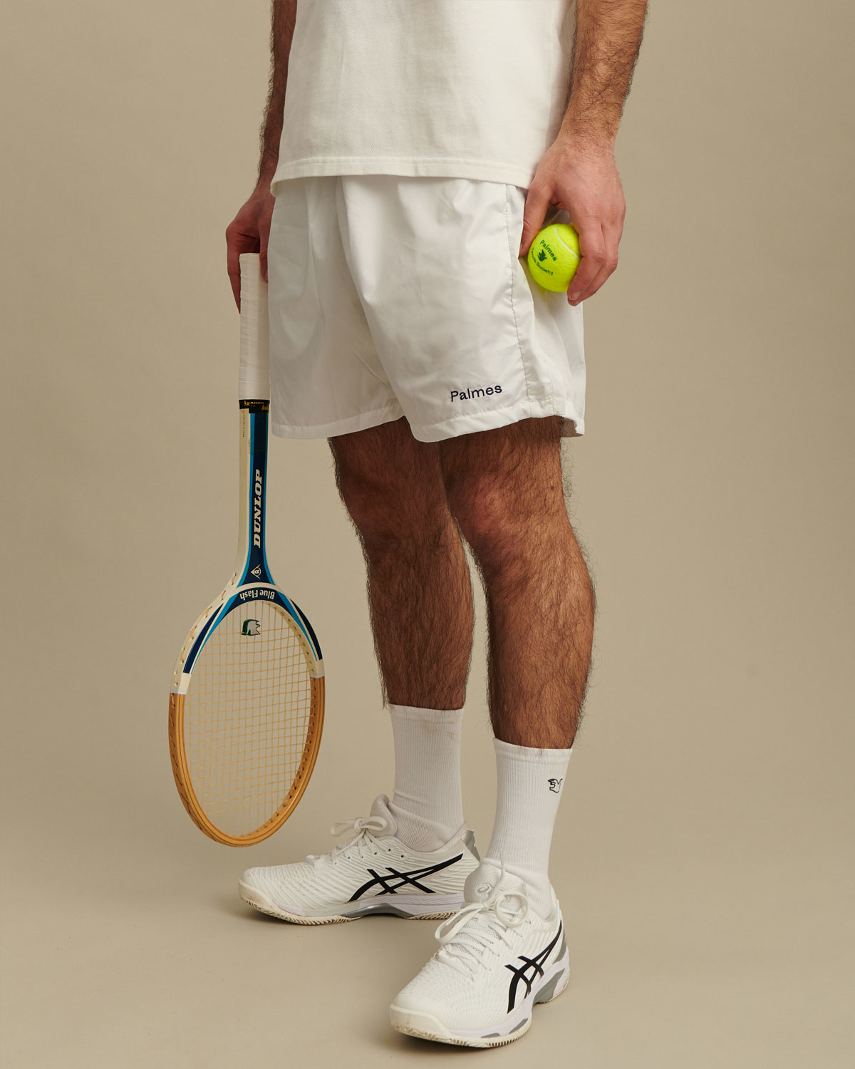 man wearing white tennis shorts while holding a yellow tennis ball and gold and blue tennis racket