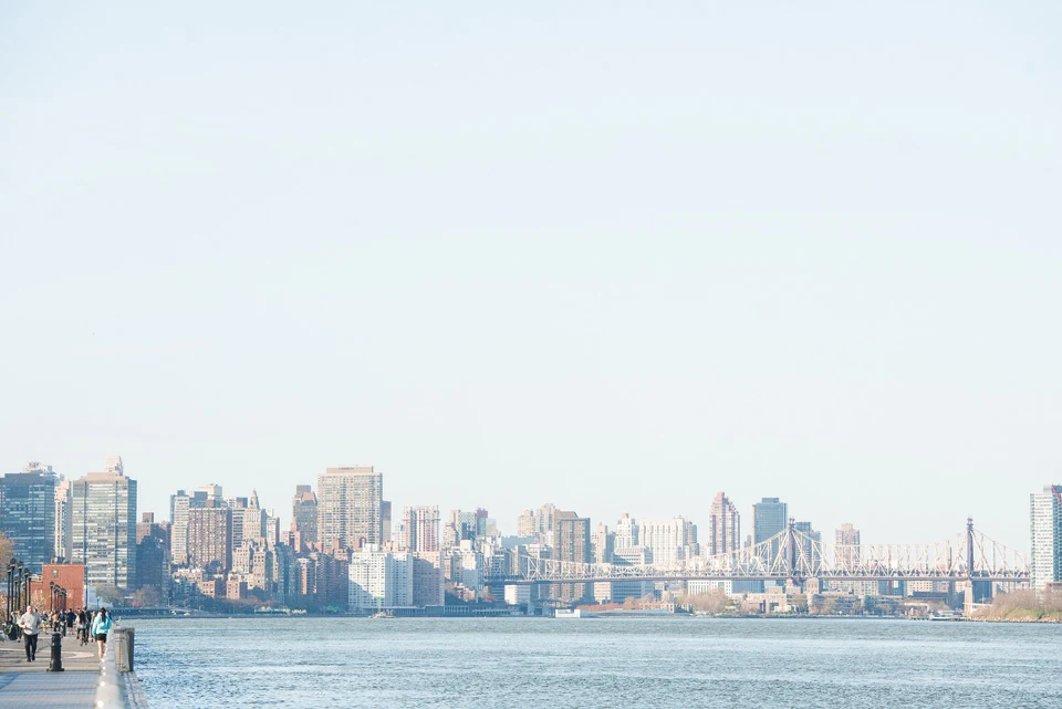 Manhattan skyline and Queensboro Bridge across the river, viewed from a waterfront walkway.