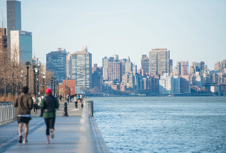 People walk along an East River promenade, with the Manhattan skyline across the water.