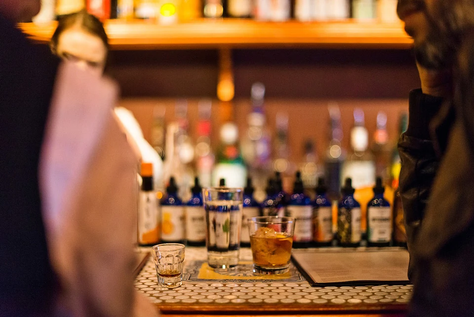 Warmly lit bar counter with liquor bottles and two drinks, patrons partially visible at the edges.