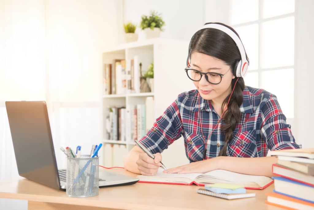 Portrait of student learning online with headphones and laptop taking notes in notebook sitting at her desk at home - mixed race Asian Chinese model