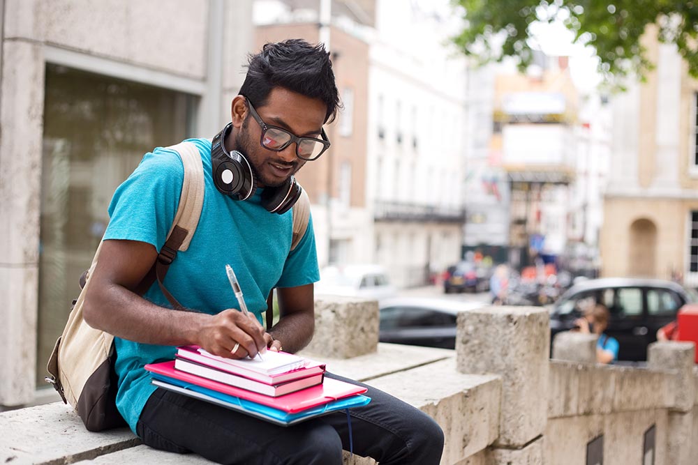 Male-student-taking-notes-at-university-outdoors