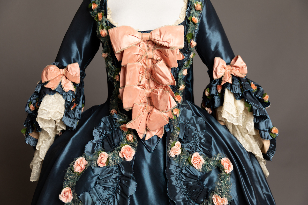 The top half of an elaborate, 18th century-style dress of dark blue material. Large pink bows create a "v" on the front of the dress, and it has rows of small pink roses along sides. Half-length sleeves end in draping blue fabric and lace.