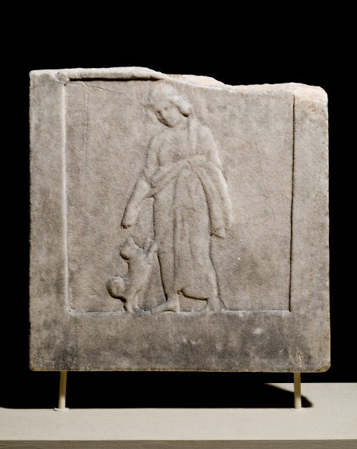 A square relief panel depicting boy who is facing forward but turns slightly as he extends his right hand toward a curly-tailed dog who leaps up at the boy's right thigh. He wears a cloak wrapped around his lower body and held in place by his left arm.