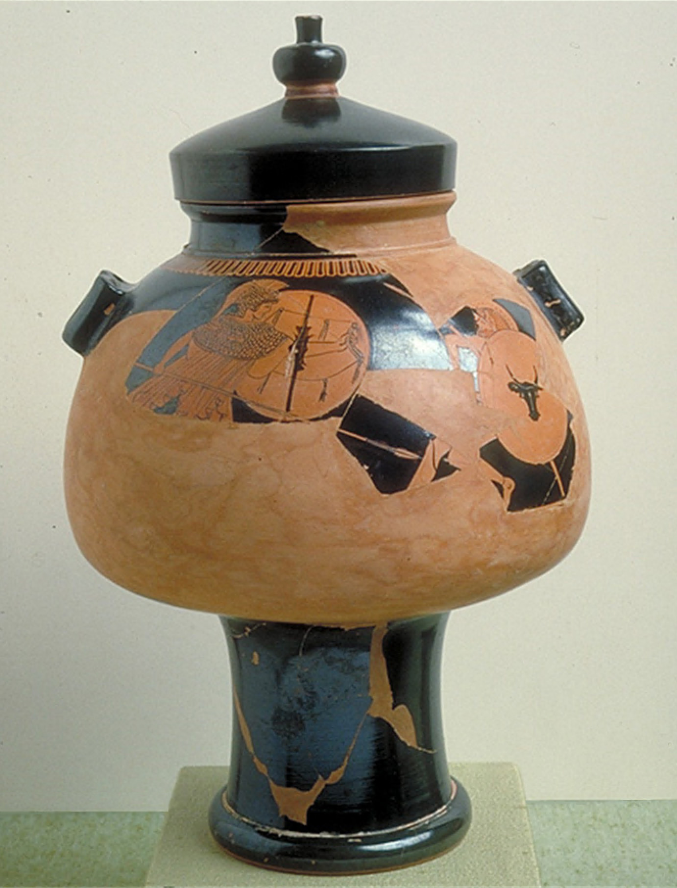 A pear-shaped pot sits on a cylindrical base. It is topped by a lid with a knob. Sections of the figural scene painted around the central area of the pot are missing. But one soldier faces another solder in battle; both are holding spears and shields. 