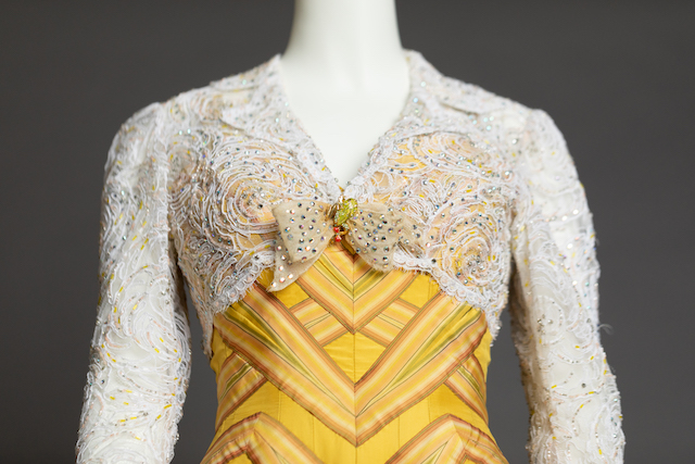 A yellow dress with orange and green stripes creating a chevron pattern is shown from the waist up. Over the dress is a white lacy bolero-style jacket decorated with rhinestones. The front of the jacket is affixed with a bow and a green frog pin.